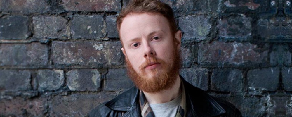 Matthew wearing a leather jacket and looking at the camera standing in front of a dark brick wall