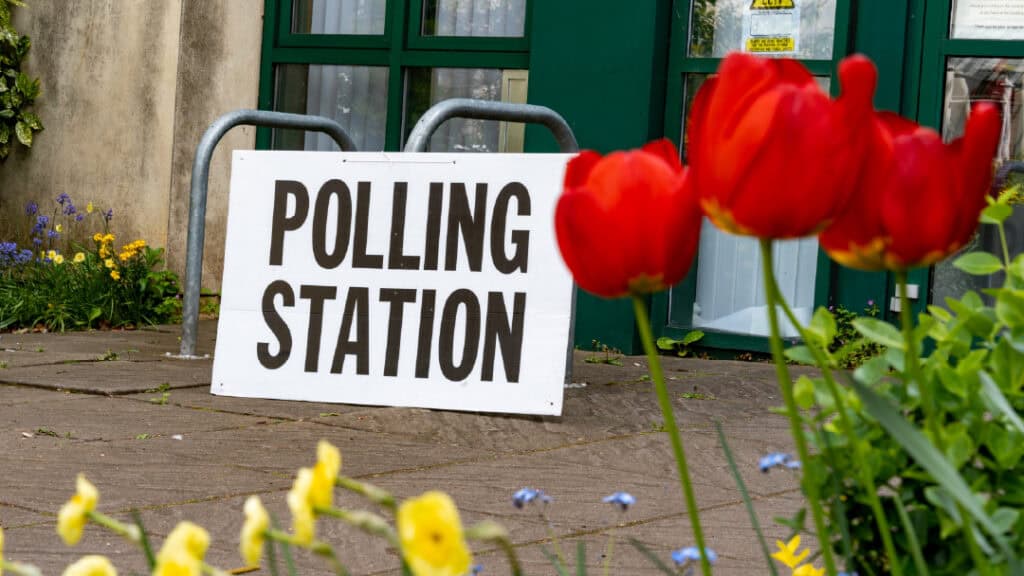 A "Polling Station" sign is displayed outside a building. Bright red tulips and yellow flowers are in the foreground.