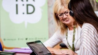 Two women sat beside each other at a table talking. One wears headphones and has a screen beside her. An RNID banner can be seen in the background.