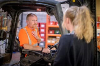 A man sitting in the driver's seat of a fork-lift truck talking to a woman who is standing with her back to the camera