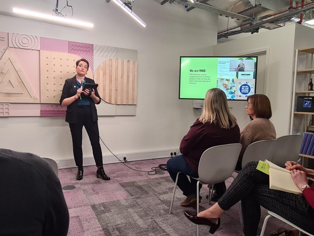 Claire Lavery, our Associate Director for Employment, stands at the front of a room giving a talk with a microphone. Employees of Google's Accessibility Discovery Centre are sat in chairs and listening to Claire.