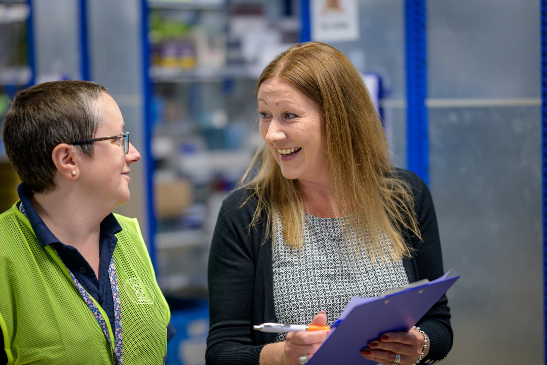 Two women talking and smiling in a public space, one has a clipboard and pen