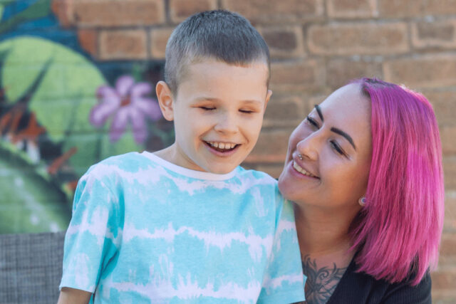 A woman with pink hair kneels down to a young boy and looks lovingly at him.