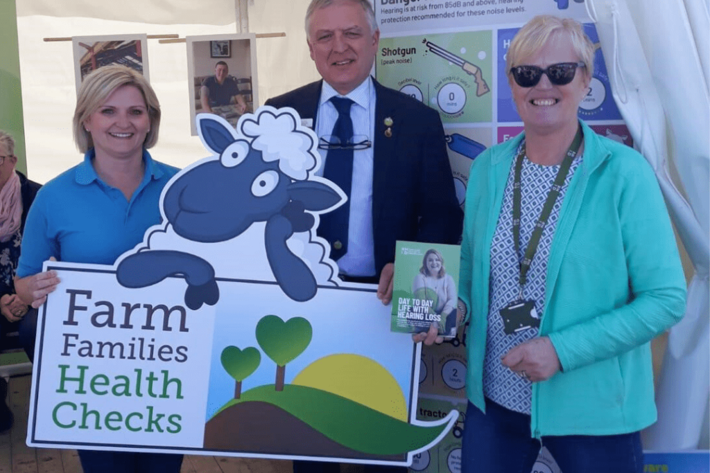 Three people smiling outside, holding a sign that says: 'Farm Families Health Checks'.