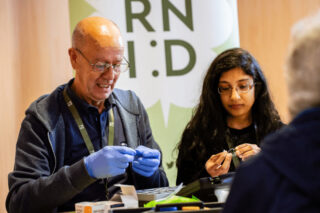 Two people at an RNID event stand, seated and handling hearing devices