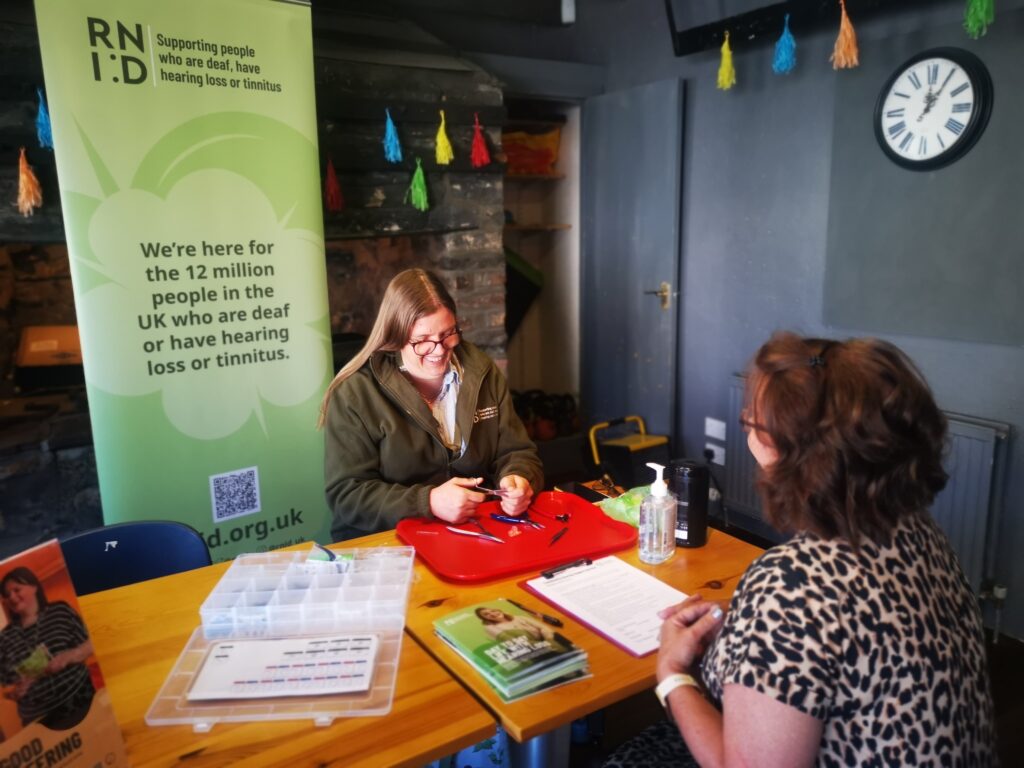 RNID volunteer Sarah in Lauder, sitting at a table and smiling as she replaces hearing aid tubes for the person sat opposite. There are RNID leaflets on the table and a banner in the background. 