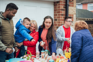 A group of people gathered outside around a table laid with cakes.