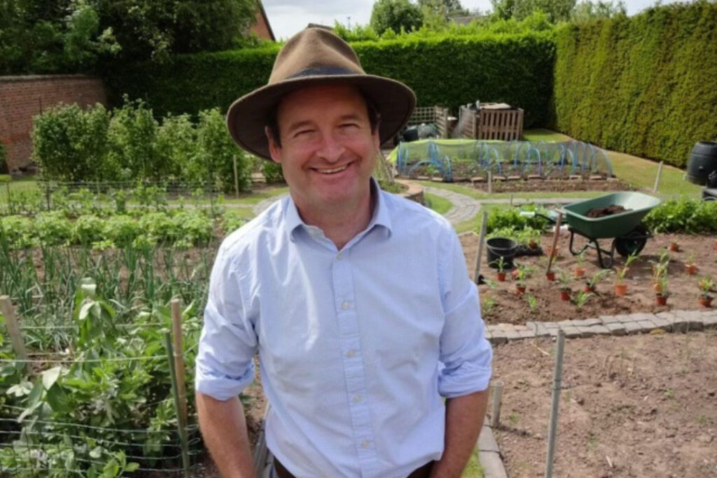 Hugh, a man in a shirt and sunhat, stands outside in a garden and smiles at the camera. 