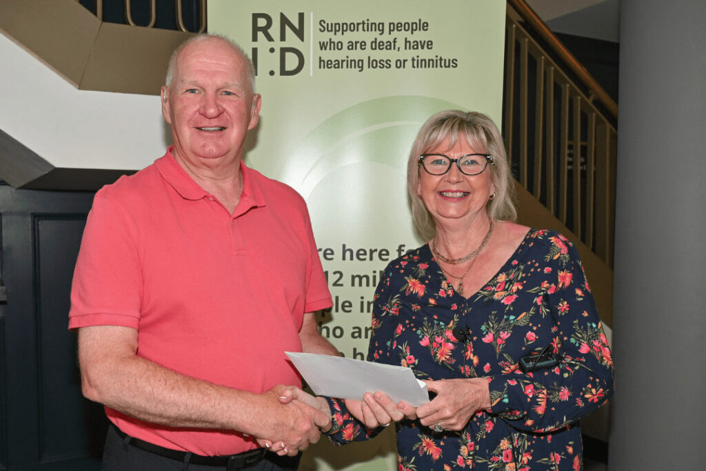 A man and woman shaking hands and smiling for a photograph, holding a certificate, in front of a RNID banner, 