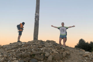 A smiling woman wearing an RNID t-shirt stands at the summit of a mountain with her hand outstretched.