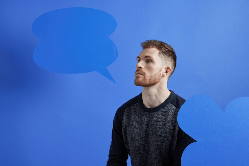 A man wearing a black jumper sits in front of a blue background. He wears a hearing aid. Icons of speech bubbles are surrounding him.