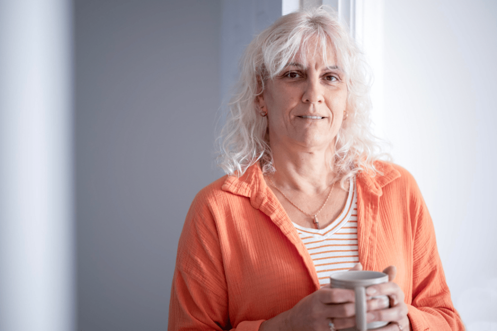 A woman in an orange shirt in front of a white wall, holding a mug of tea, and making eye contact with the camera. 