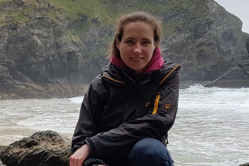 Researcher Dr Magdalana Zak wears a raincoat and sits on a rock beside the sea.