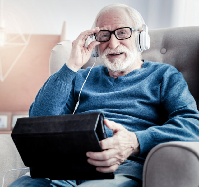 Elderley man seated in an armchair, wearing glasses and headphones, holding a tablet device and smiling at the camera.