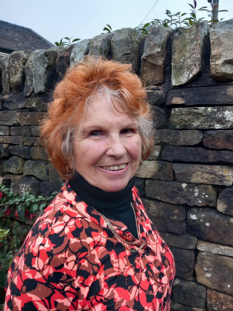Linda, an RNID support service user, stands outside by a dry stone wall, smiling at the camera.