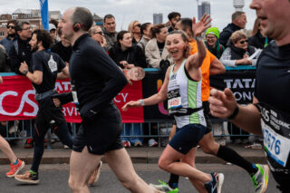 People running in the London marathon, a women runner laughing and waving at the camera. A crowd behind the runners are cheering.