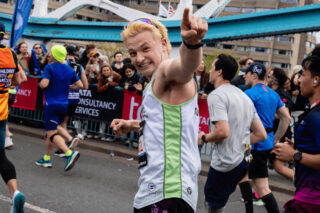 A man taking part in a running event smiling at the camera and pointing. A crowd cheers in the background