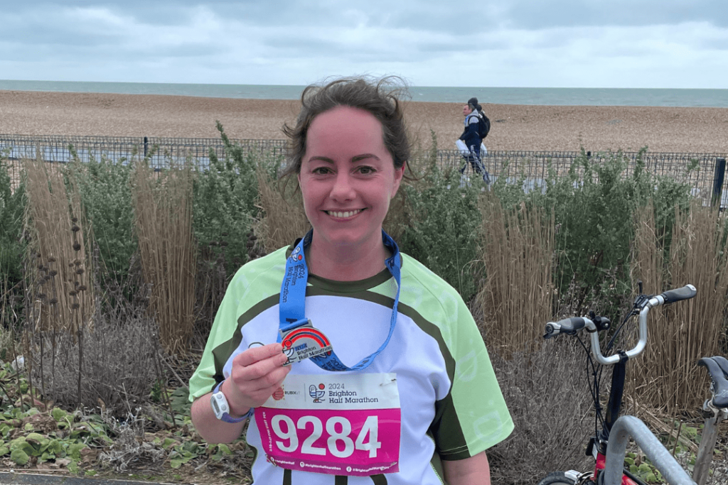Katie Thomas in a RNID running tshirt shows her medal, smiling at the camera. She stands in front of the Brighton coast. The sea is in the background. 