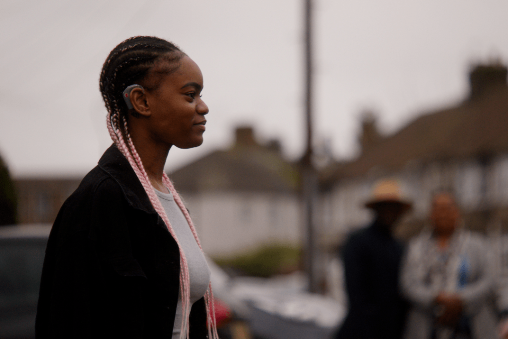 Sarah stands outside, facing sideways to the camera. Her assistive hearing technology is visible at her ear. Her pink braids are over her shoulders. Her parents stand in the background, blurred.