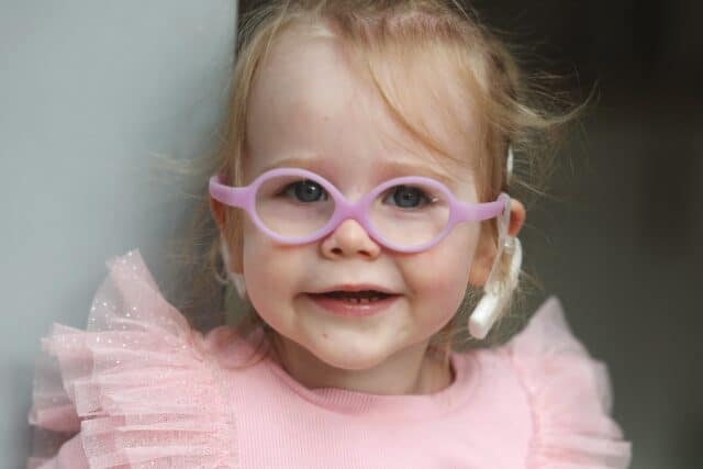 A photo of Tilly, a young child in a pink dress and glasses, wearing a cochlear implant. She's smiling and looking into the camera.