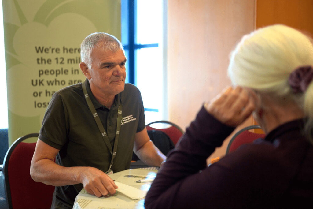 An RNID volunteer seated at a table talking to a woman who has their back to the camera