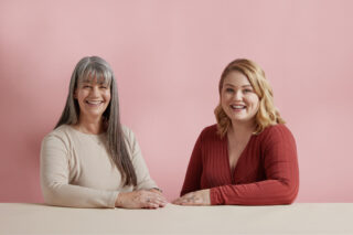 A photograph of two women sitting at a table, smiling at the camera. They are both hearing aid users.