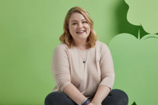 A smiling woman sitting with folded legs on the floor against green background.