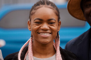 Sarah, a deaf BSL user. She stands outside in front of a blue car and smiles at the camera. Her hair is in pink braids. She wears a cochlear implant.