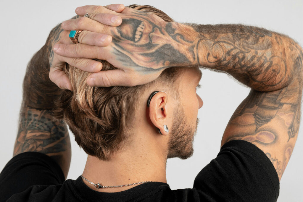 A man faces away from the camera with his hands interlaced on the back of his head. His arms are covered in tattoos. He wears a diamond stud and a hearing aid.