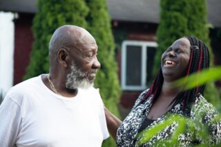 Two people standing in a garden. The man is older and looking at the younger woman, who laughs at something he has said.