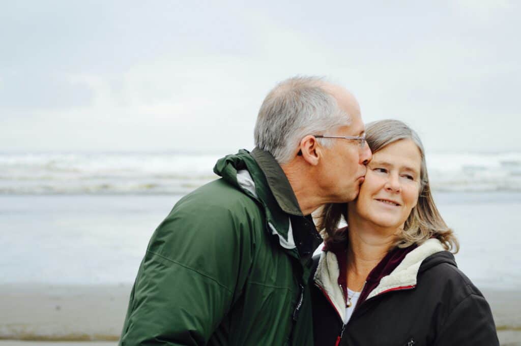 An older man and woman standing by the sea. The man kisses the woman on the cheek.