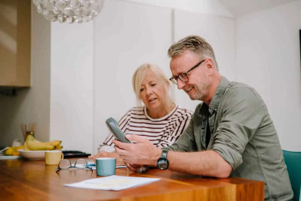 A couple seated at their kitchen table looking at their landline phone.