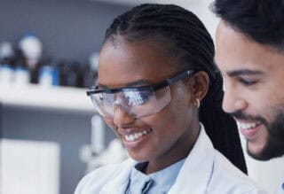 Two scientists working in the lab, wearing goggles and white coats.