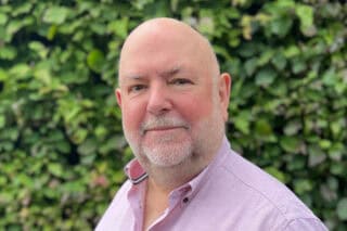 David Evans Connevans, who wears a pink shirt and has a bald head, stands in front of greenery.
