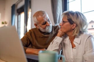 A man and woman sitting at a dining table, looking online, via laptop.