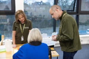 Two RNID volunteers helping someone with their hearing aid at an RNID drop-in session.