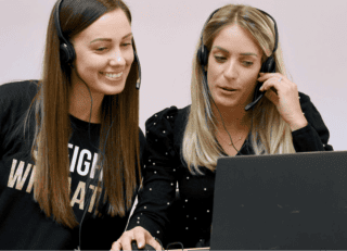 Two women look at a laptop screen, smiling. They both wear headsets.