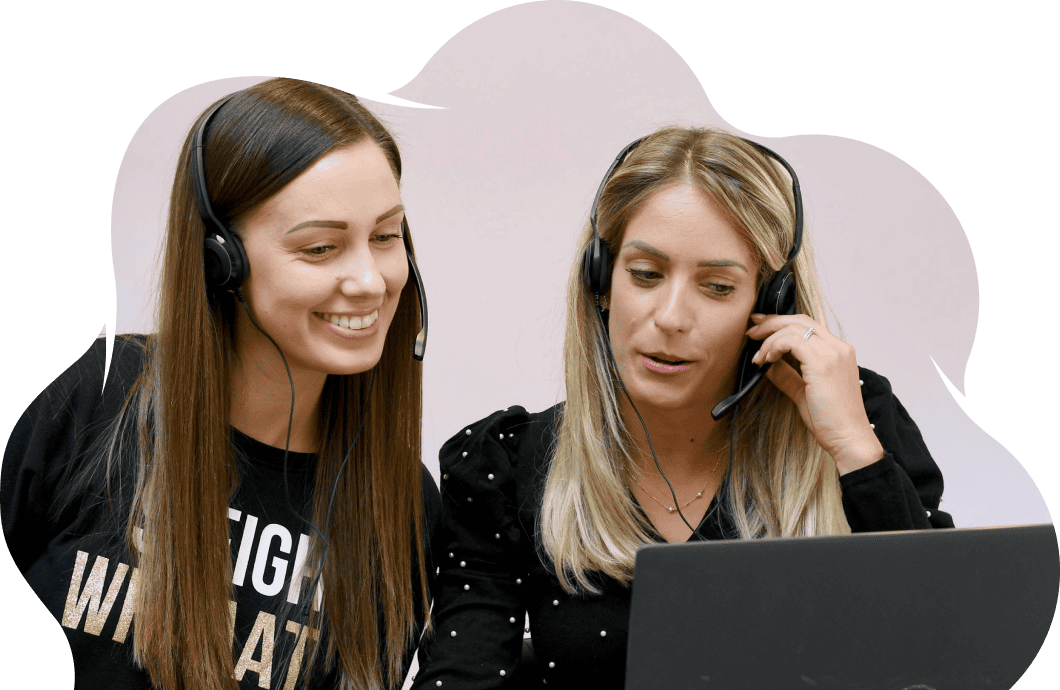 Two women look at a laptop screen, smiling. They both wear headsets.