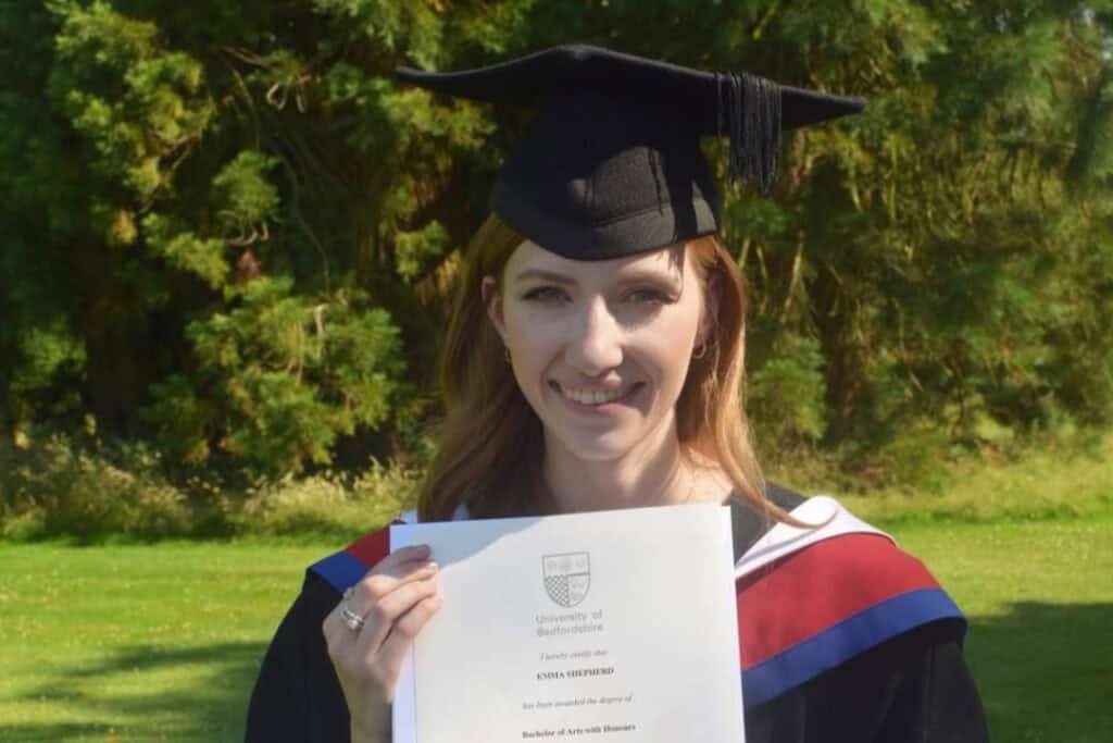 A smiling woman wearing a graduation gown and cap holds a certificate proudly.