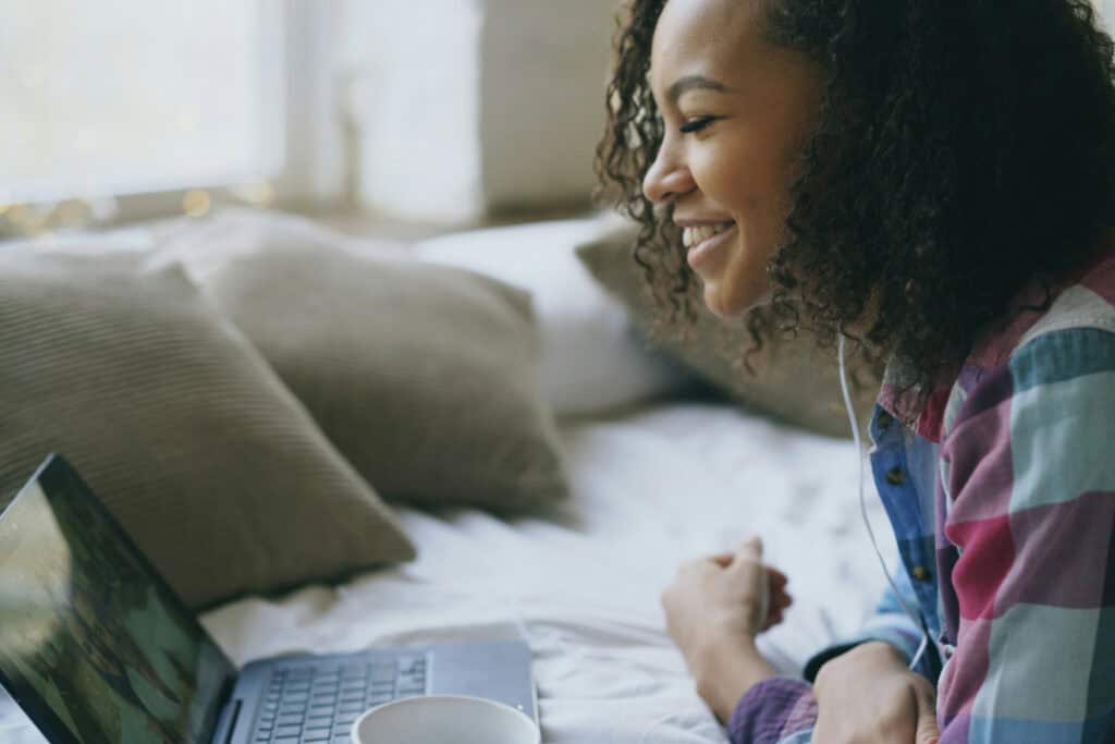 A woman lies on her bed and is smiling at her laptop. 