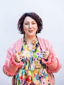 A smiling woman with brown hair wearing a colourful dress puts both thumbs up.
