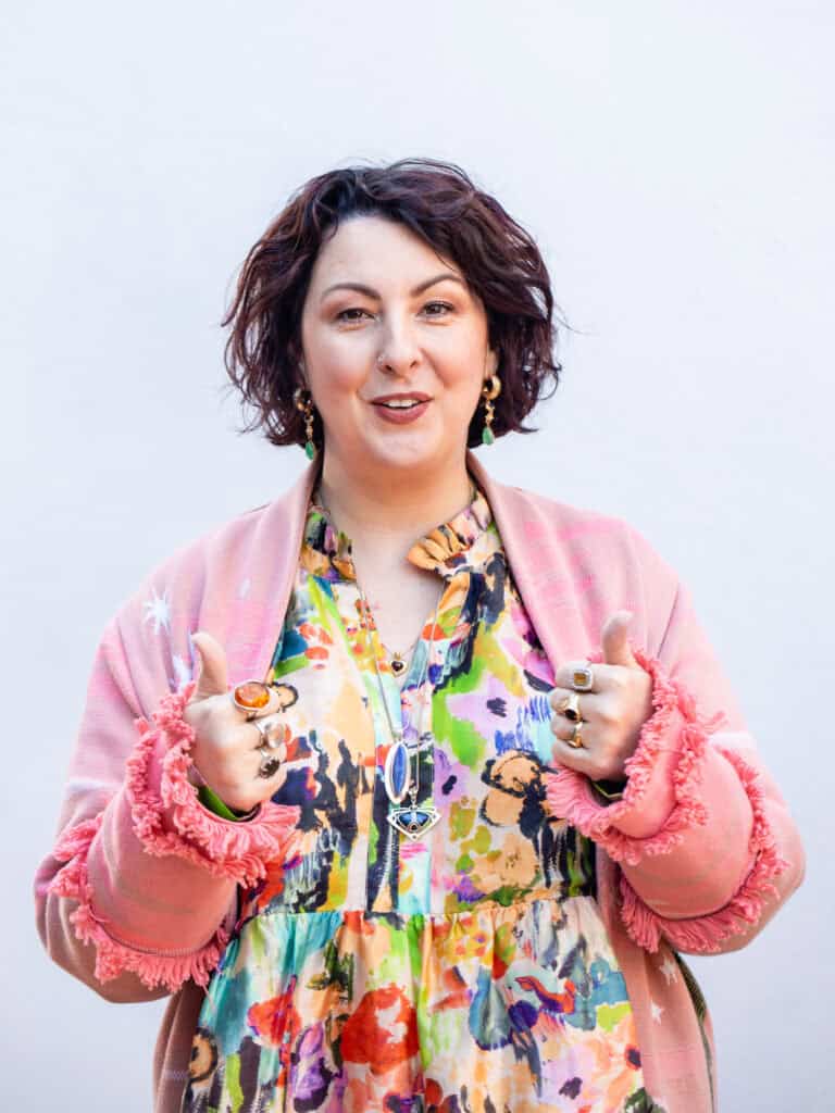 A smiling woman with brown hair wearing a colourful dress puts both thumbs up.