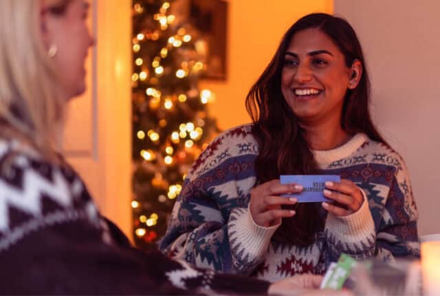 Two women have fun playing RNID's Christmas Dinner Game. They are in a dimly lit room with a Christmas tree twinkling in the background.