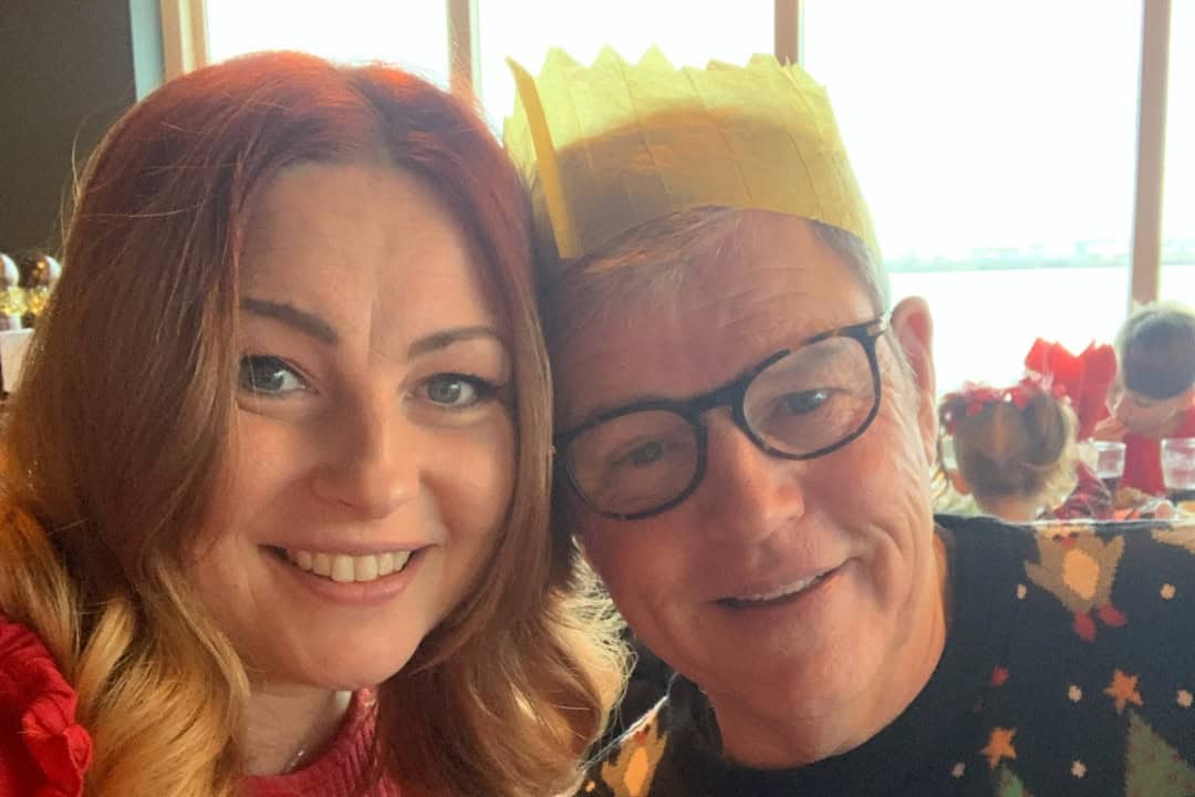 A smiling father and daughter. They are dressed festively, with the father wearing a yellow paper Christmas hat and festive jumper and the daughter wearing a red jumper.