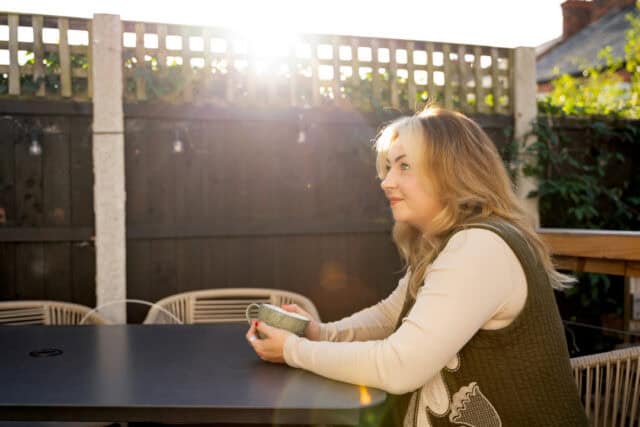 RNID supporter, Kate, sits in a garden, holding a mug. Sunlight streams through the garden fence.