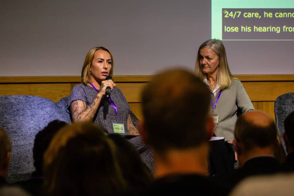 A woman sits on a stage and talks into a microphone in front of an audience at RNID's Hearing Therapeutics Summit 2025.