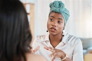 A black woman with a blue headscarf on talks animatedly to someone.