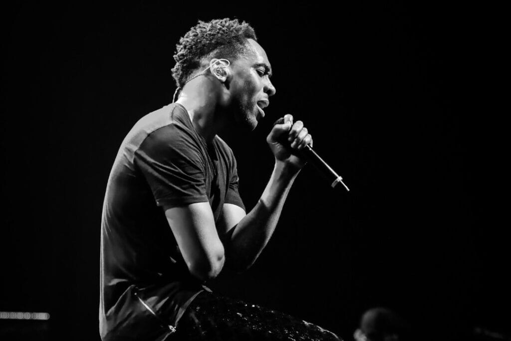 A black and white photo of pop star, Lemar, sat on stage, singing into a microphone. Photo credit: Christie Goodwin