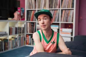 Matt sits on the floor, leaning on a sofa. He is in a room with shelves of records, and he wears a cap and sports vest.