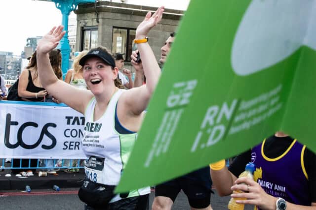 A woman runs London Marathon wearing an RNID running vest. She waves and smiles.